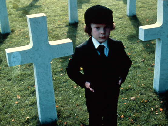 “The Omen,” the 1976 classic horror film that imagines an ambassador’s son as the antichrist - here, Damian stands in a black suit and tie among white crosses in a cemetery.