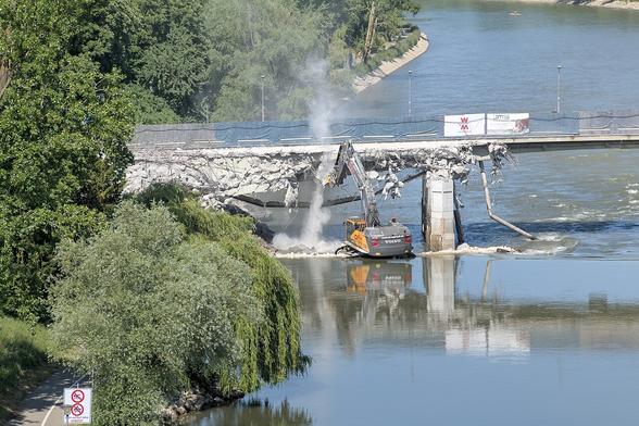 Aus erhöhter Perspektive ein Blick auf die Donau mit dem Rest der Gänstorbrücke. Im Vordergrund steht ein gelb-grauer Raupenbagger in der Donau und pulverisiert die von links kommenden schon zerbröselten Reste der westlichen Gänstorbrücke. Im Hintergrund ist die noch erhaltene Ostseite der Brücke zu sehen, über die der Verkehr geleitet wird.