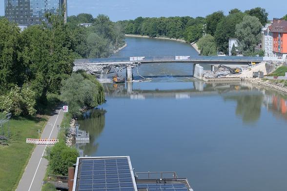 Vom selben erhöhten Standort wie im vorigen Bild, aber in der Totalen: Die Donau verläuft längs nach hinten, von links nach rechts ist die noch erhaltene Ostseite der Brücke zu sehen. Jeweils links und rechts zwischen den Behelfspfeilern und dem Ufer stehen je ein Bagger auf Aufschüttungen in der Donau und zerkleinern die zerbröselten Reste der Westseite der Brücke.