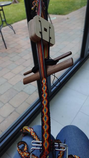A tablet woven band of many colours in the foreground, with a background of floor tiles, window and exterior brick paving.  The band is on a backstrap loom with a wooden band shuttle placed in the shed of the loom. The tablets are made of white cardboard. The further end of the loom is out of the picture and is tied to the window latch.  The nearer end is wrapped round a metal band lock which is looped over an out of shot waist belt (backstrap).