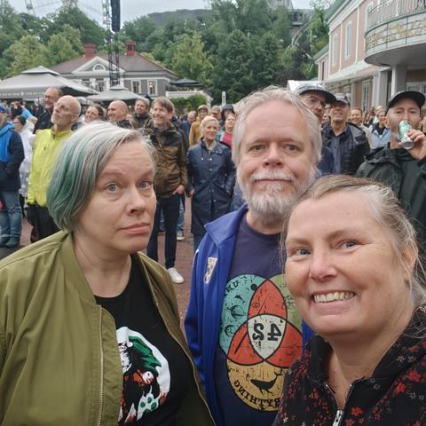Three white middle aged people. To the left a woman with grey hair with green streaks and a green bomber jacket. In the middle a man with white hair and white beard, a nerdy t-shirt and a blue athletic jacket, to the right mr, looking silly: a broad, smiling face, my hair in a bun, and a black corduroy anorak with red flowers.