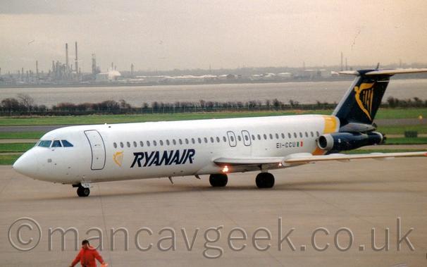 Side view of a twin engined jet airliner taxiing from right to left.
The plane is mostly white, with dark blue "RyanAir" titles on the lower forward fuselage next to a yellow, winged Irish harp.
The tail is dark blue with a larger version of that yellow Irish harp logo, and a yellow band wrapped around the rear fuselage.
The dark blue engines are mounted on the sides of the rear fuselage, just under the tail.
The black registration "EI-CCU" are on the lower rear fuselage, just ahead of the engines.
Grey concrete apron fills the foreground, with a man in a red jacket walking out of frame at the bottom left.
In the background, green grass leads up to a wide river, with an industrial landscape on the far side, under grim grey sky.