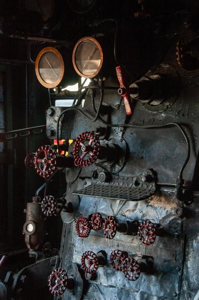 Dark, chiaroscuro view of an old steam locomotive control panel. There are many valve handles painted dull red, some illegible gauges, piping, and other complicated-looking bits of unknown purpose.