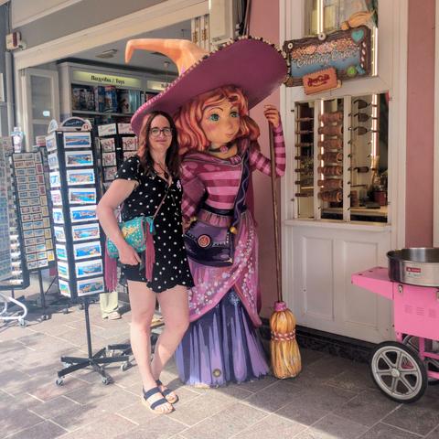 Posing with a store mascot. She's a witch in pink and red striped top, red hair, a pink and purple skirt, a massive orange and pink witch's hat, and a broom. She's got a purple cross-body handbag and is standing in a jaunty pose. I'm also a witch, albeit undercover, in a black minidress with white polka dots, Labradorite pendant, red hair, but sadly no witch's hat nor broom. I've got a turquoise and dusky pink cross-body handbag and am standing in a jaunty pose.