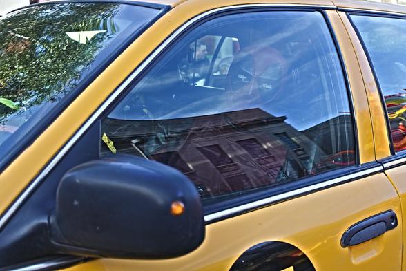 The image captures a close-up, eye-level view of the passenger side of a bright yellow vehicle, which appears to be a taxi. Through the slightly tinted window, a reflection of a brick building with several windows is visible, suggesting an urban setting. Inside the vehicle, behind the window's reflection, the distinct red and black masked head of Deadpool is clearly visible, facing towards the viewer. The sunlight catches the upper part of the car and the reflection, creating a mix of light and shadow on the scene. The car's black side mirror with an orange indicator light is prominent in the foreground, extending from the side of the vehicle.
