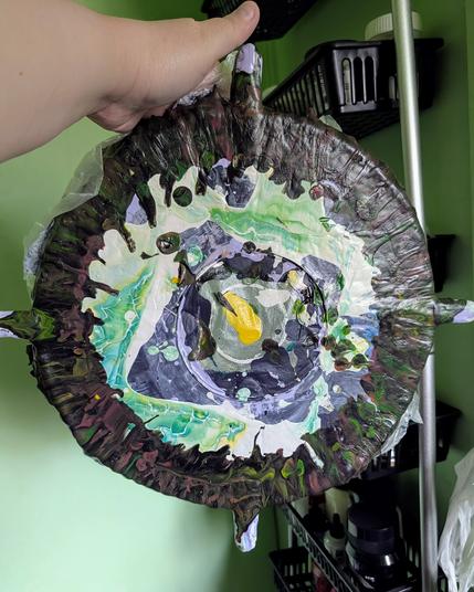 My hand holding up my soon-to-be-discarded circular canvas spinner cover. You can see various layers and colors from some of the different fluid art projects I've worked on more recently. There are four popsicle sticks poking out of different sides that are also covered in paint.