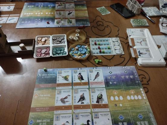Partida a dos jugadores del juego de mesa Wingspan. Se ve el despliegue completo con los tanleros de jugador,las cartas, las fichas y dados de alimento y los objetivos de ronda.