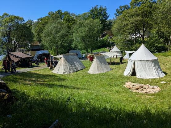 A group of white canvas tents arranged on grass near a few green trees, with several people in medieval-style clothing standing and walking among the tents. In the background, a clear blue sky are visible.