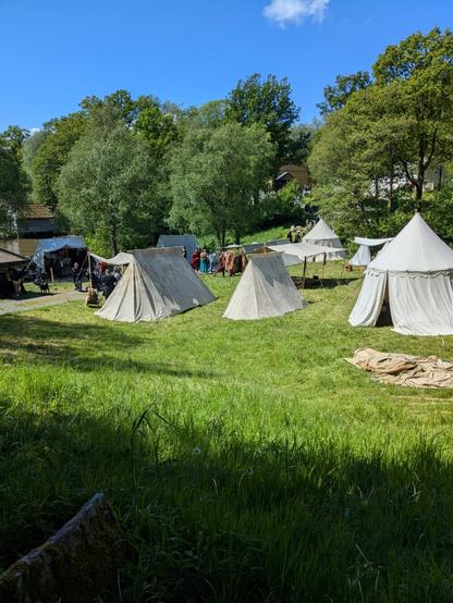 A group of white canvas tents arranged on grass near a few green trees, with several people in medieval-style clothing standing and walking among the tents. In the background, a clear blue sky are visible.