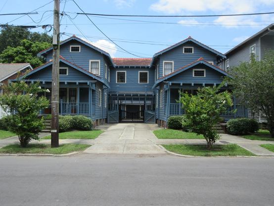 Photo of  pair of matching wooden houses of the "camelback" style common in late 19th century New Orleans residential districts - single story up front and 2 story behind. In this case the two houses are joined on the upstairs midway back on the property, with a driveway between the house fronts and a gateway under the connecting section.  The structure is symmetrical and painted dark blue.