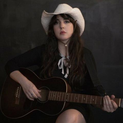 A white woman with long dark hair sits on a stool with crossed legs and a black acoustic guitar. She is wearing a black dress and white cowboy hat and is seated against a black backdrop. She looks directly at the camera.
