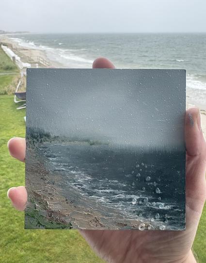 Photo of a hand holding an original seascape oil painting by Tisha Mark., "Yarmouth, MA, Cape Cod Plein Air Painting" 4"x4" oil on Ampersand Gessobord (2025). Rainy day seascape painting with a dark blue sea meeting a beachy coastline on the left side of the painting. The location where the painting was created is visible in the background.