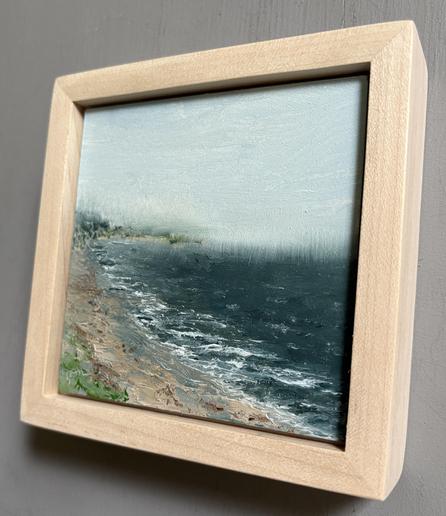 Original seascape oil painting by Tisha Mark, "Yarmouth, MA, Cape Cod Plein Air Painting" 4"x4" oil on Ampersand Gessobord (2025), shown in a maple frame at an angle from right to show painting texture and depth of frame. Rainy day seascape painting with a dark blue sea meeting a beachy coastline on the left side of the painting.