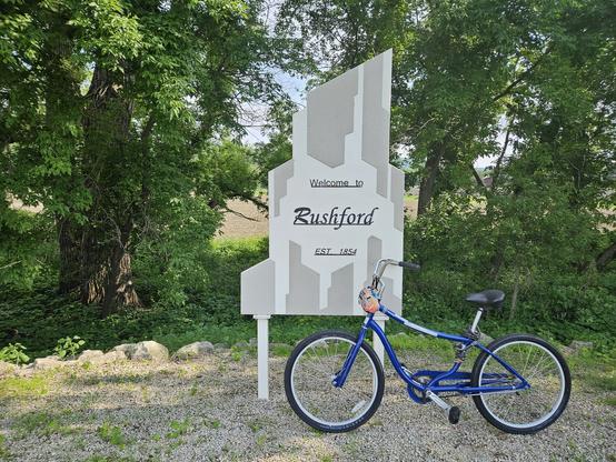A blue bicycle leans against a white and grey sign. The sign says "Welcome to Rushford est. 1854. The bicycle is on grey gravel with patches of green grass . Behind are bushes and  trees with green leaves.