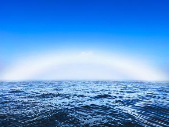 A serene ocean view under a clear blue sky, with a faint rainbow arc visible in the distance above the water. The surface of the sea has gentle waves.