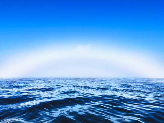 A serene ocean scene with gentle waves under a clear blue sky. A faint rainbow arching over the horizon adds a touch of color to the tranquil atmosphere.
