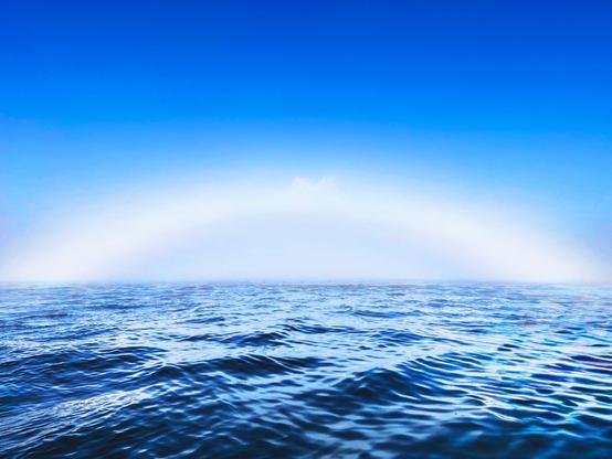 A smooth ocean surface under a clear blue sky, with a faint mist creating a soft arc on the horizon.