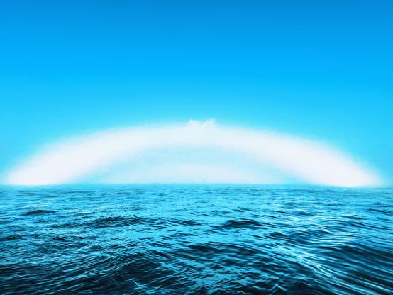 A vibrant blue ocean under a clear sky, featuring a soft white arc that resembles a distant rainbow or mist. The water appears calm with gentle waves.