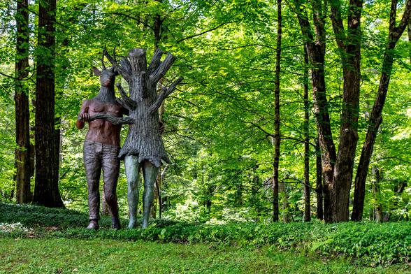 In a very green forest, two figures (presumably a sculpture). One figure has the body of a man and the head of a deer with antlers. The second figure has human legs, but the upper torso is a tree stump with no head.