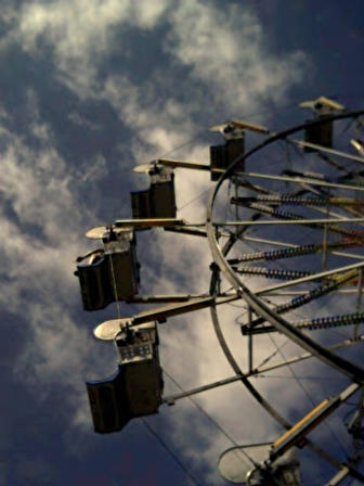 Looking through a Ferris wheel (Most of which is out of the frame on the right) up into a blue sky with clouds.