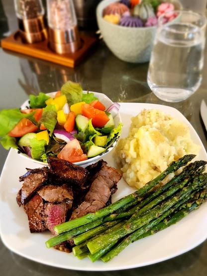 A white plate with steak, mashed potatoes, asparagus, and a bowl of fresh garden salad.