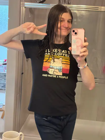 A tgirl flashes the peace sign wearing a black tshirt that says "I like cats and coffee and like 3 people"