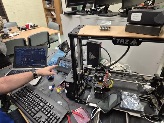 A desk piled with a laptop, keyboard and a disassembled 3D printer. Finger pointing at an interface screen for the 3D printer