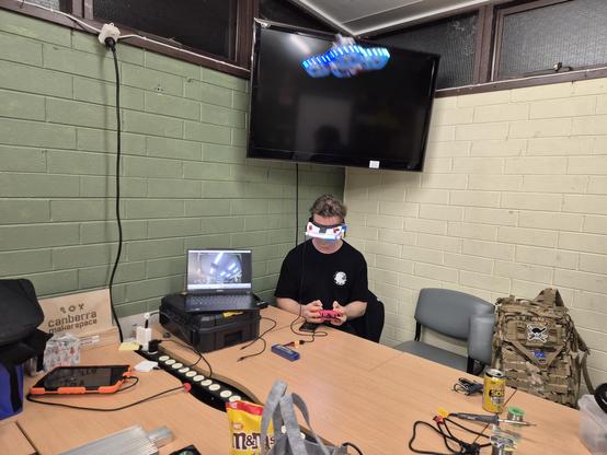 Person sitting at table piloting an FPV drone. The drone is captured mid-air.