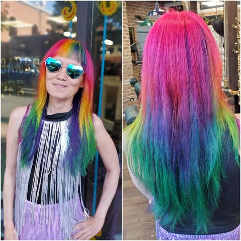 Split-panel medium shot. Left side shows a waist-up view of an Asian woman(me) with rainbow-colored shoulder-length straight hair, rainbow ombre bangs, wearing heart-shaped blue mirrored sunglasses is smiling at the viewer. She's wearing a black tank top with a silver fringe shirt and a purple sequin pants. The background is slightly blurred, showing a storefront with "BARBERS" signage and a colorful swirl decoration. The right side is a back-facing medium shot showcasing her rainbow ombre hair from pink at the roots to green at the tips. Background is a brick wall and hanging lamps, in what seems to be a barbershop. The composition focuses on showcasing the colorful and vibrant hair dye effect.