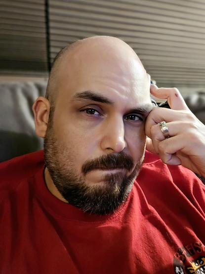 Matt B is sitting indoors on a couch, wearing a red shirt. He is resting his head on one hand, with his index finger against his temple and a ring visible on his ring finger. His expression appears thoughtful or slightly weary. Horizontal blinds are visible in the background.