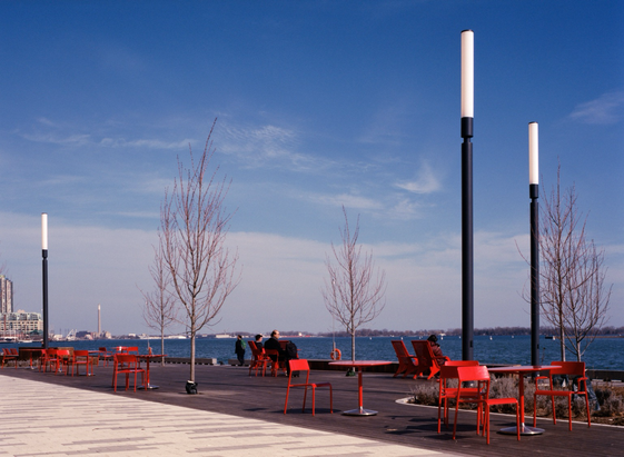 A public space with a wooden deck and bright red chairs and tables built on an industrial quay. The trees are bare and the harbour stretches across the view.