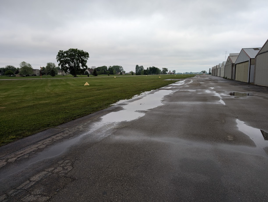 Outdoor picture of a rain soaked apron at an airport. Clouds overhead and hangars are visible going off into the distance. The grass runway is cut nicely but given the puddles on the apron it is definitely hiding some water.