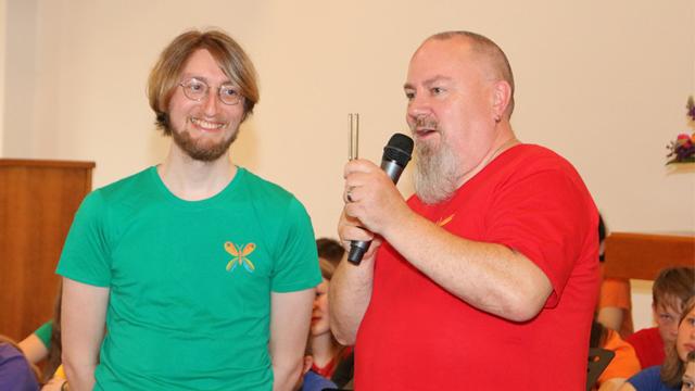 Rechts steht Pastor Lutz Brückner. Ein Mann mit sehr kurzem Haar und langem Bart in rotem T-Shirt mit der rechten Hand ein Mikrofon haltend und in der linken eine Stimmgabel. Links daneben steht Johannes Tunger. Ein Mann mit Vollbart und Brille in grünem T-Shirt, der seinen Nachbarn ansieht.