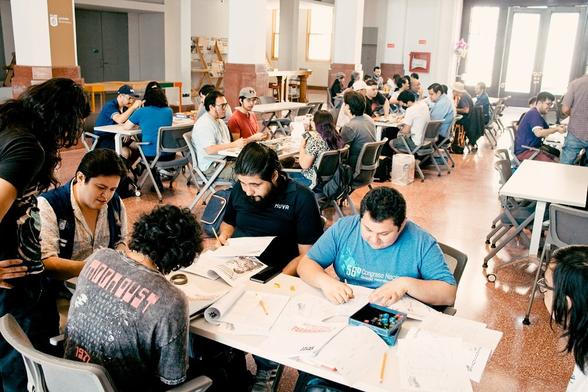 En primer plano se ve una mesa con 5 personas sentadas llenando hojas de personaje antes de iniciar una partida de un juego de rol. Al fondo se observan un montón de mesas con personas sentadas alrededor de ellas y jugando distintos juegos.