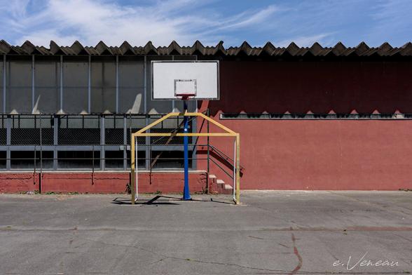 A sports field with a basketball board. Behind the wall of an ochre gym with a small staircase for access.
