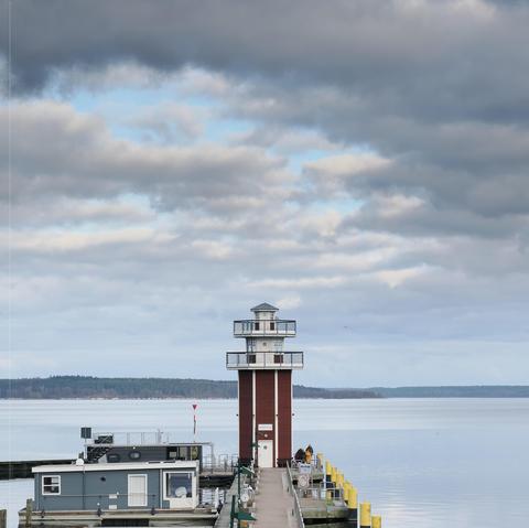 Lighthouse centered on a wooden pier extending into a calm body of water, flanked by a small building to the left. Two individuals stand on the pier near the lighthouse. Cloudy sky with patches of blue overhead, distant tree line visible on the horizon.