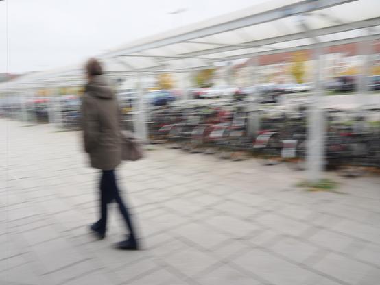 Blurred image of a person wearing a hooded coat and walking past a row of bicycles under a metal canopy. The bicycles are parked in a designated area, and there are autumn-colored trees in the background. The ground is paved with gray tiles, and the overall scene has a motion blur effect.