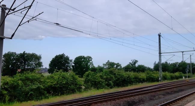 Foto an einer Zugstrecke. Am Himmel eine scharf gezogene Grenze zwischen Wolken und blauem Himmel.