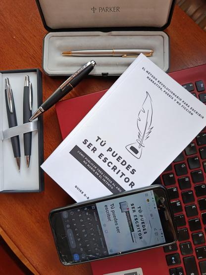 El libro «Tú puedes ser escritor», de Quike D-B, al lado de un teclado de ordenador portátil, varios bolígrafos de marca y un móvil con la pantalla encendida con un teclado táctil y el título del libro.