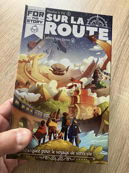 Boîte du jeu de cartes narratif « Sur la route ». L’illustration aux couleurs chaleureuses représente un groupe de voyageureuses toustes différent·e·s et pas forcément humain·e·s de dos au premier plan, qui regarde au loin toutes les aventures possibles en train, en bateau, en van, en fusée, en dirigeable, à dos de dromadaire… Si on revient au premier plan, on remarque un chat roux aux pieds du groupe, qui se retourne vers nous.