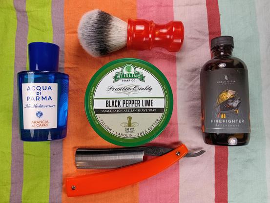 (Le Chat Mistral description)

The image showcases a collection of grooming items arranged on a striped background. At the top left, there's a blue bottle of Acqua di Parma cologne. To the right of the cologne, there's a shaving brush with a red handle and white bristles. Below the brush, centrally located, is a green tin of Stirling Soap Co. Black Pepper Lime shaving soap. To the right of the soap, there's a brown bottle of Noble Otter Firefighter aftershave. At the bottom of the image, there's an orange-handled straight razor. The items are neatly arranged, suggesting a traditional shaving setup.
