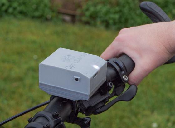 Ein kleiner grauer Kasten, 3D gedruckt, auf einem Fahrradlenker, links ein kleiner Knopf. Rechts hält eine Hand den Lenker fest.