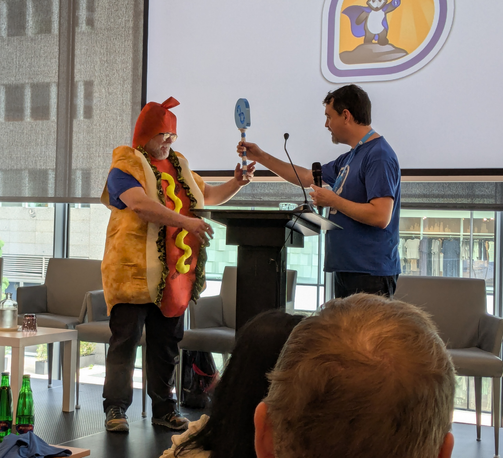 Matthew in a blue t-shirt handing over the Fedora sceptre (3d printed) to Jeff who is wearing a hotdog costume.
