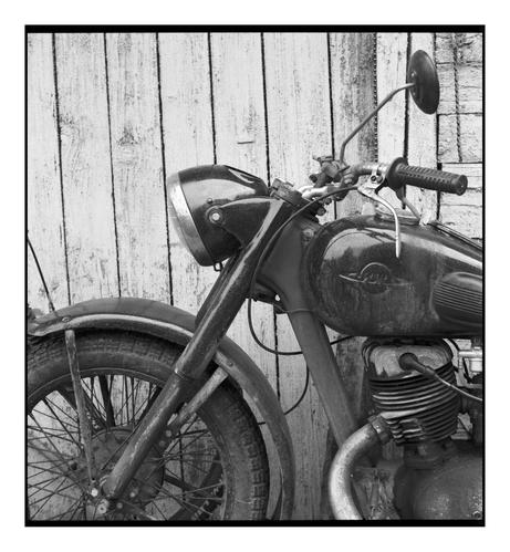 Close-up of an old Soviet motorcycle parked in front of a weathered wooden fence. The image shows the front wheel, fuel tank, and handlebars, highlighting age and texture. Shot on Kodak TMAX 400 film with a Yashica Mat-124 G medium format camera.