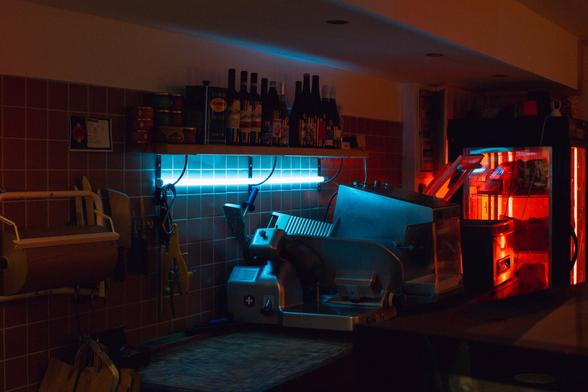 Interior of a butcher shop. A metallic slicing machine is under a cold blue neon light, while the background is in the shadows, lit by a bright red neon light