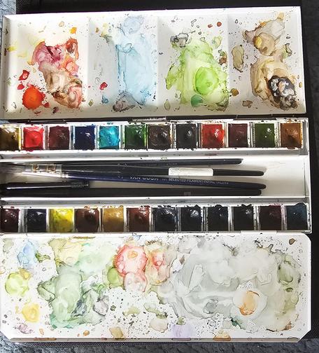 Aquarellkasten mit 24 Farbnäpfchen und 2 ausgeklappten Mischflächen, über und über mit Farbresten. In der Mitte sind diverse Pinsel erkennbar.