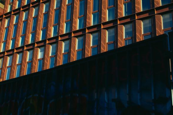 In the background, a brightly sunlit building, with a very angular architecture made of straight lines. Rust-colored  metal is framing vertical, narrow windows repeating over and over. In the foreground, slicing the building diagonally, is a backlit construction fence, made of metal, with paintings and graffitis. There is a high contrast between the golden hour light on the building and the shadows on the fence.
