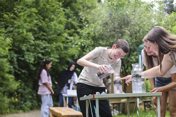 Drei Studentinnen stehen an einem Tisch in einem Garten. Eine füllt Schlamm in eine Filterflasche. Die anderen beiden schauen zu. Eine zeigt auf die Flasche. Der Tisch steht in einem Garten. Im Hintergrund dichtes Grün und weitere Studentinnen.  Foto: Fabry/PHKA