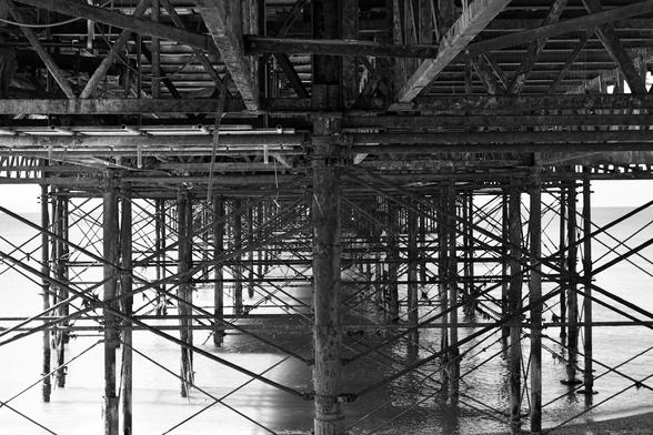 Monochrome photo taken from the beach of the substructure of Brighton Palace Pier. First constructed 1891-1901, designed by R St George Moore. Grade II* listed. Madeira Drive, City of Brighton & Hove, UK.