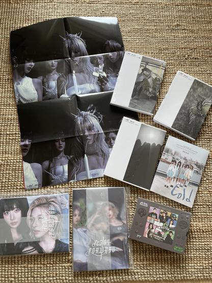 The image features a collection of music albums and merchandise, including a folded poster displaying four individuals with distinct hairstyles, several album covers in plain white packaging, and a portable gaming device. The items are arranged on a woven surface.